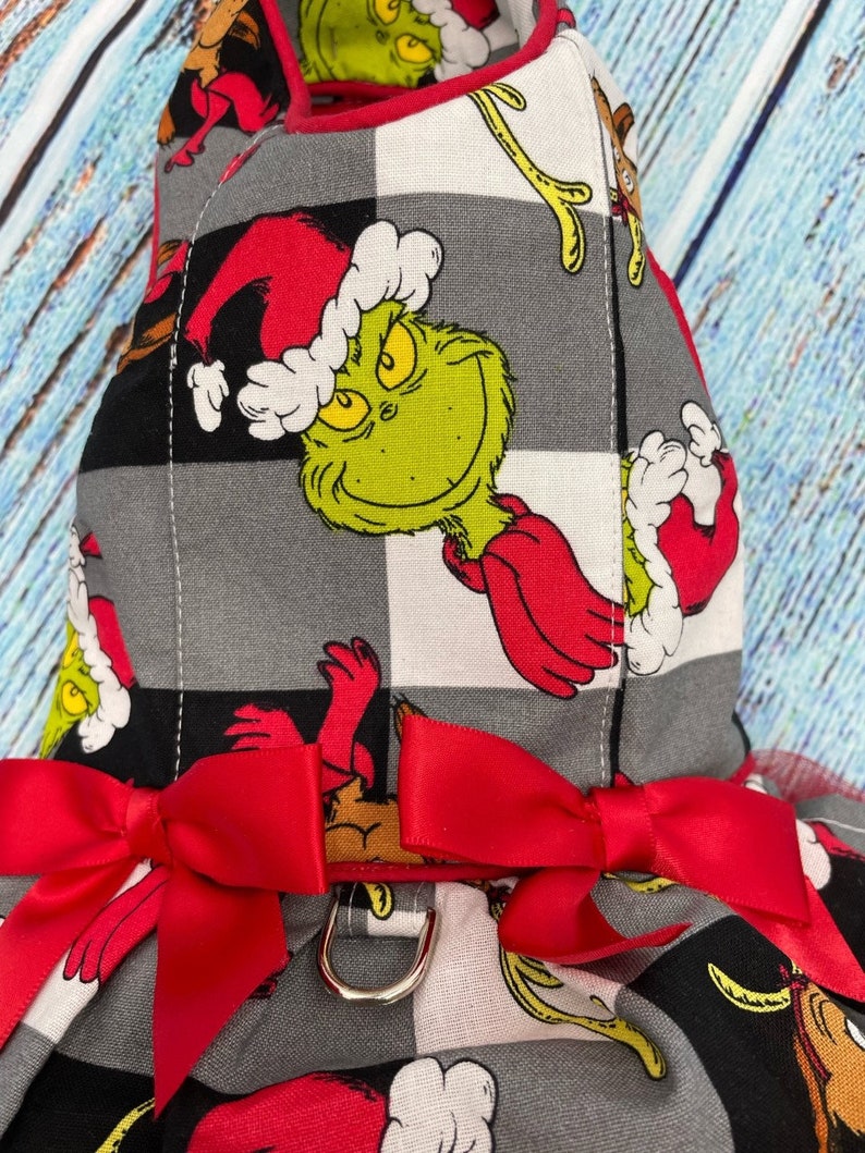 May include: A pet harness featuring a grey and black checkered pattern with a repeating print of a green character wearing a red Santa hat. The harness includes red fabric trim, two red satin bows at the front, and a silver metal D-ring.