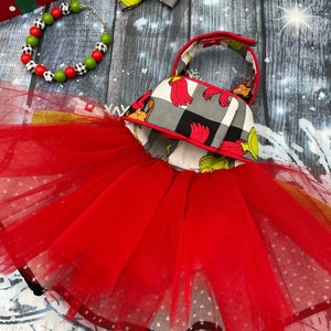 May include: A red tulle tutu dress with a bodice featuring a grey and black buffalo plaid pattern and green character illustrations. The set includes a matching hair bow and a beaded bracelet with red, green, and black and white chequered beads.