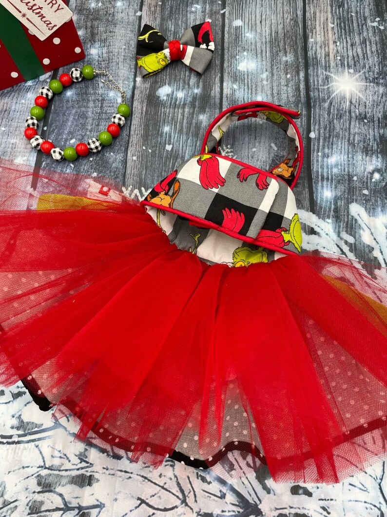 May include: A red tulle tutu dress with a bodice featuring a grey and black buffalo plaid pattern and green character illustrations. The set includes a matching hair bow and a beaded bracelet with red, green, and black and white chequered beads.