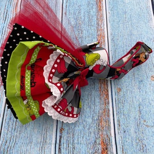 May include: A layered fabric accessory featuring a red tulle overlay, black and white polka dot fabric, and a red panel with green character illustrations and the text Merry Grinchmas. It includes white lace trim, a red ribbon bow, and a strap.