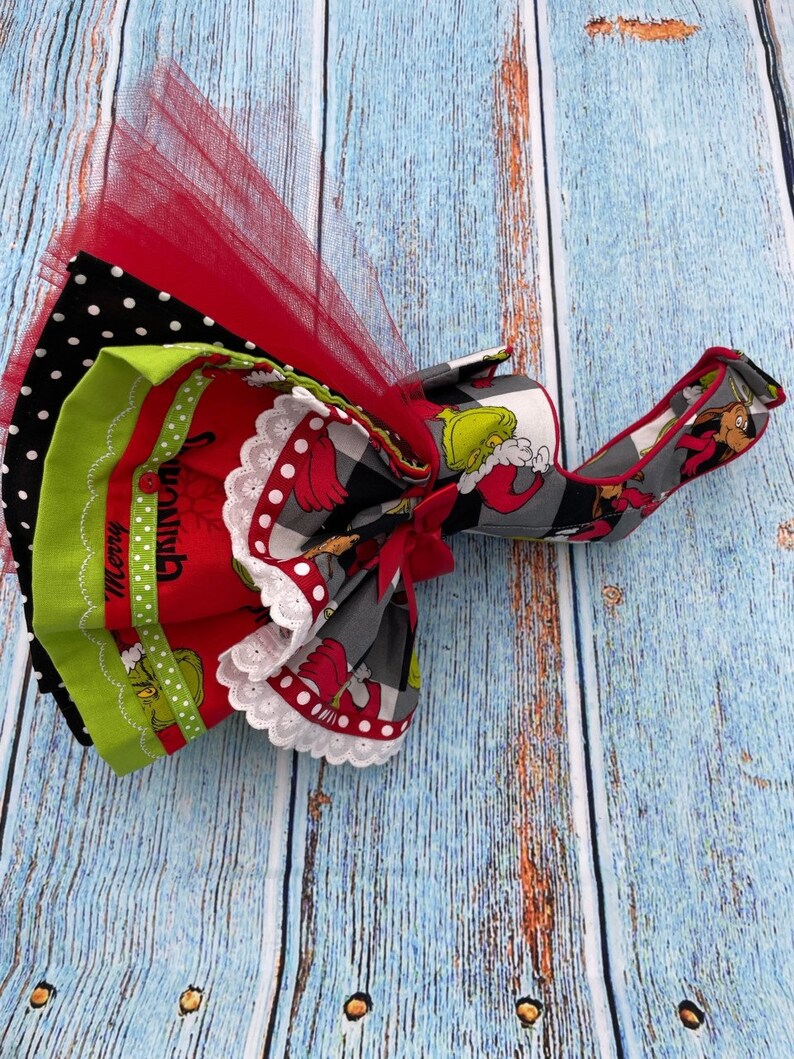 May include: A layered fabric accessory featuring a red tulle overlay, black and white polka dot fabric, and a red panel with green character illustrations and the text Merry Grinchmas. It includes white lace trim, a red ribbon bow, and a strap.