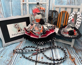 May include: A Halloween-themed pet apparel display featuring a dress, hat, and leash. The dress has a gray bodice with orange and black accents, a black and white checkered skirt, and a black bow. The hat is black and white with a spider. A black and white leash is also included.