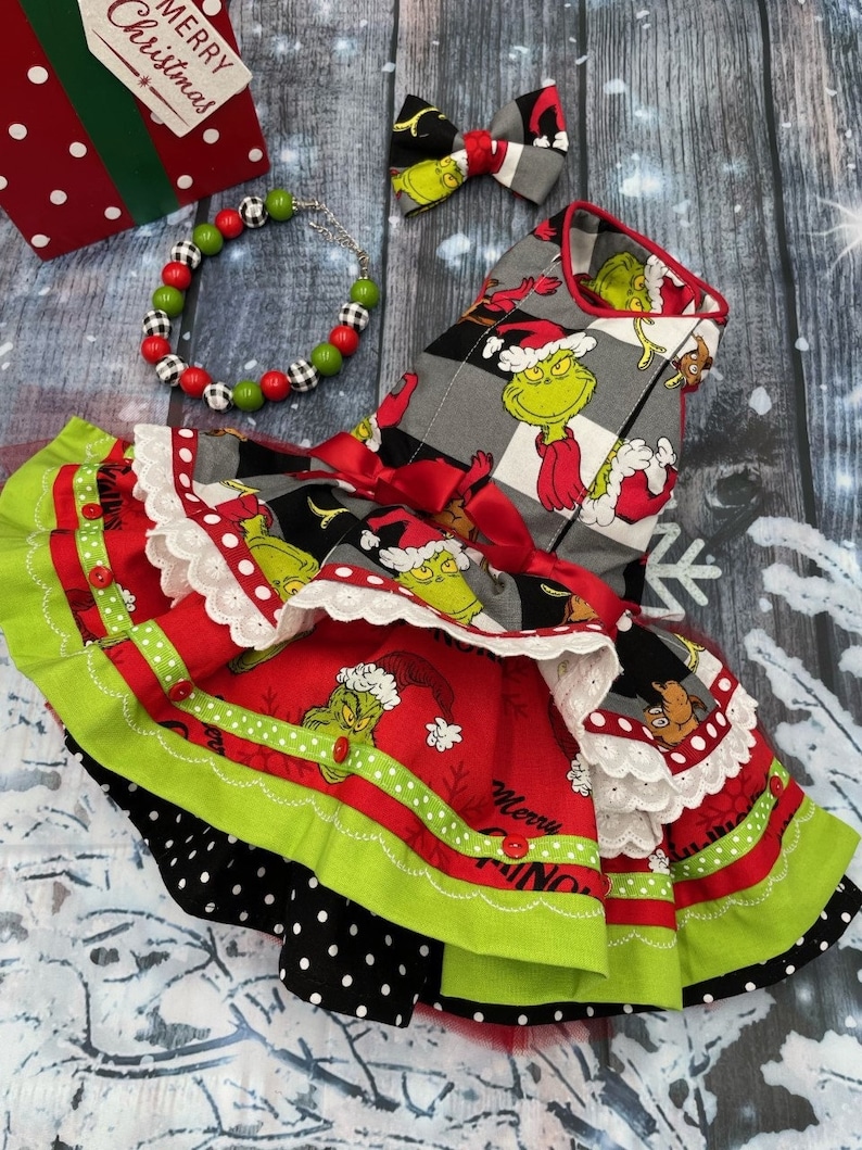 May include: A pet dress featuring a Grinch print on a grey and black checkered bodice and a red skirt with white lace trim, green ruffles, and red buttons. Includes a matching bow tie and a beaded necklace with red, green, and black-and-white beads.