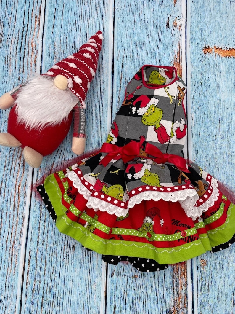May include: A gnome with a red and white striped hat and a white beard next to a dress featuring a Grinch print on a grey and black buffalo plaid bodice, a red skirt with green trim, white lace accents, and a red ribbon bow at the waist.