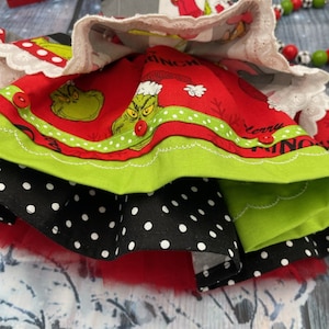 May include: A multi-layered skirt featuring a red fabric layer with a Grinch print, the text Merry Grinchmas, and small red buttons. It includes a black layer with white polka dots, a solid lime green layer, and a white lace-trimmed top layer.