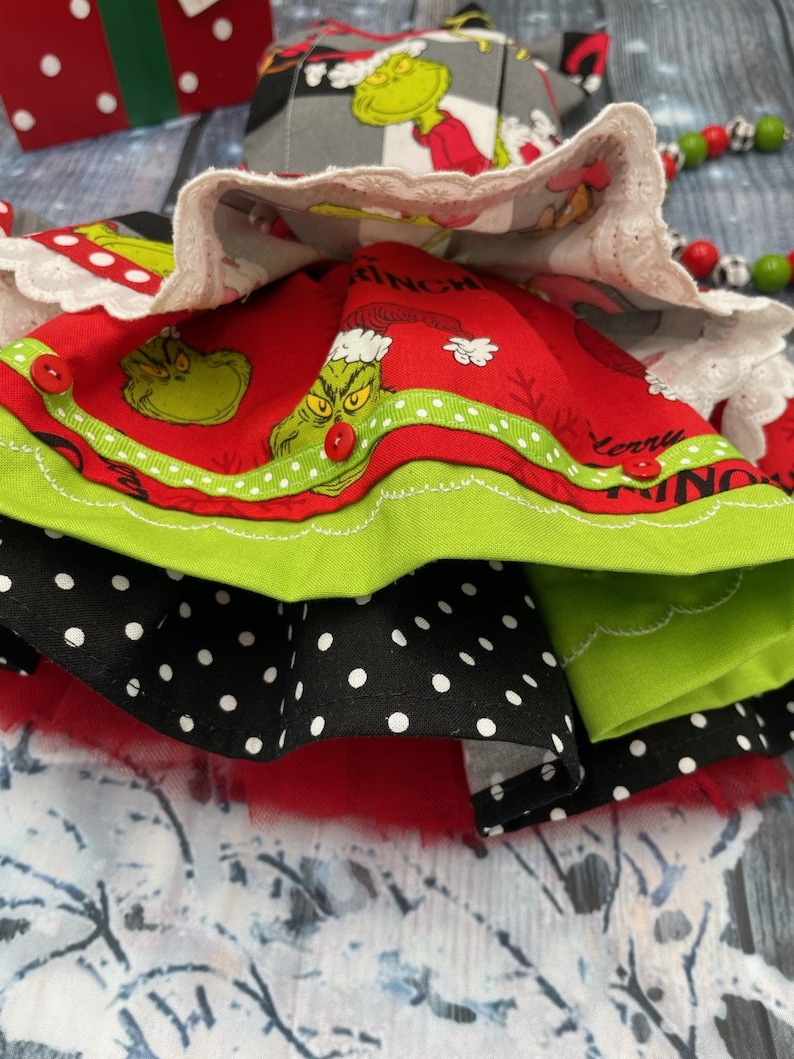 May include: A multi-layered skirt featuring a red fabric layer with a Grinch print, the text Merry Grinchmas, and small red buttons. It includes a black layer with white polka dots, a solid lime green layer, and a white lace-trimmed top layer.