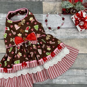 May include: A brown pet dress featuring a gingerbread man, candy cane, and Christmas tree print. The dress has a red bow at the waist, pink trim, and a layered skirt with white eyelet lace and red and white vertical stripes. A beaded bracelet is nearby.