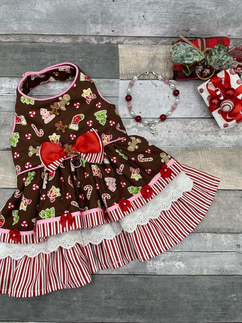 May include: A brown pet dress featuring a gingerbread man, candy cane, and Christmas tree print. The dress has a red bow at the waist, pink trim, and a layered skirt with white eyelet lace and red and white vertical stripes. A beaded bracelet is nearby.