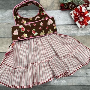May include: A brown bib-style apron featuring a festive print of gingerbread men, Christmas trees, and candy canes with pink trim. The bottom section consists of a gathered skirt with a red and white vertical stripe pattern and a ruffled hem.