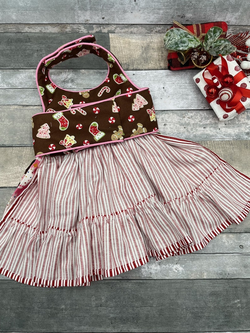 May include: A brown bib-style apron featuring a festive print of gingerbread men, Christmas trees, and candy canes with pink trim. The bottom section consists of a gathered skirt with a red and white vertical stripe pattern and a ruffled hem.