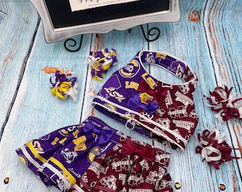 May include: Purple and maroon dog skirt with LSU and Mississippi State logos. The skirt has a white ribbon trim and a purple bow. The skirt is paired with a matching purple and maroon dog harness with a white ribbon trim and a purple bow.