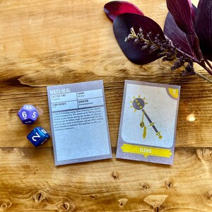 May include: Two dice, one blue with the numbers 6 and 5, and one green with the number 2, are on a wooden surface.  A grey card with the text "Mass Heal" and the text "Casting Time 1 action" is on the left.  A grey card with the text "Cleric" and the number 9 is on the right.  The card shows a black and white illustration of a mace with a spiked ball on the end.
