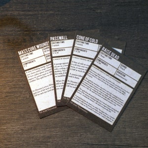 May include: Five black cards with white text describing different spells. The spells are "Mass Cure Wounds", "Passwall", "Cone of Cold", "Raise Dead", and "Cure Wounds".