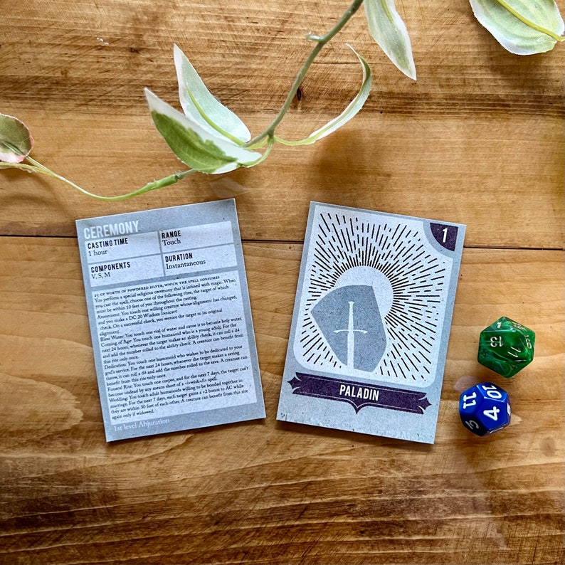 May include: A close-up of two cards, one with the text "CEREMONY" and the other with the text "PALADIN". The cards are on a wooden surface with green and blue dice.