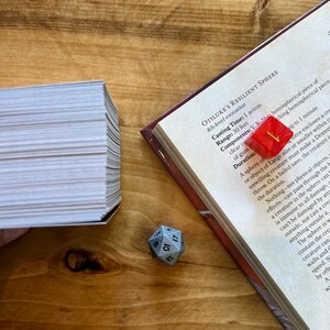 May include: A close-up of a book open to a page with the title "Otiluke's Resilient Sphere" and a description of the spell. A red d20 die is on top of the book, and a stack of white cards is in the background.