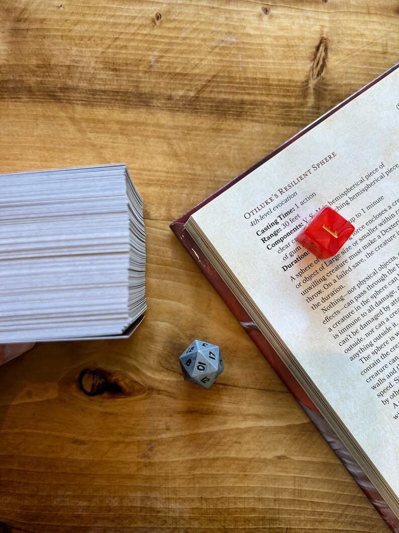 May include: A close-up of a book open to a page with the title "Otiluke's Resilient Sphere" and a description of the spell. A red d20 die is on top of the book, and a stack of white cards is in the background.