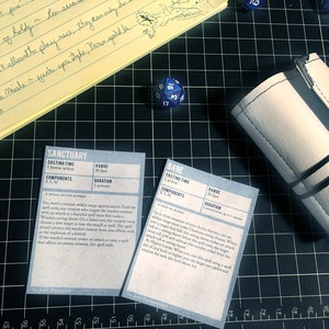 May include: Two white cards with black text on a black grid background. The cards are titled "Sanctuary" and "Bane" and contain text describing the spells. A blue 20-sided die and a white leather dice bag are also visible.