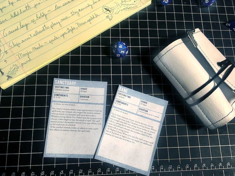 May include: Two white cards with black text on a black grid background. The cards are titled "Sanctuary" and "Bane" and contain text describing the spells. A blue 20-sided die and a white leather dice bag are also visible.