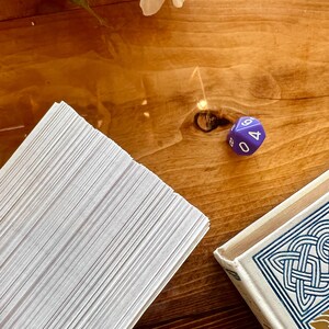 May include: A stack of white cards on a wooden table with a purple six-sided die with the number 4 facing up. The die is on the table next to a book with a blue and white patterned cover.