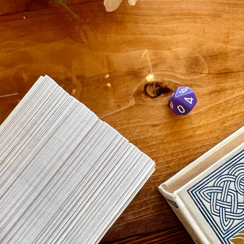 May include: A stack of white cards on a wooden table with a purple six-sided die with the number 4 facing up. The die is on the table next to a book with a blue and white patterned cover.
