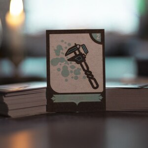 May include: A card game card with a brown background and a light brown border. The card has a number "2" in the top right corner. The card has a blue and black illustration of a wrench. The card has the word "ADJUSTER" in the bottom center.