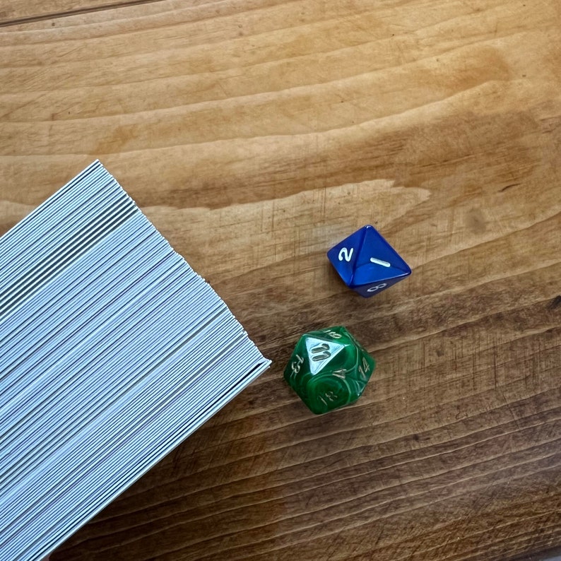 May include: A stack of white cards with a blue and green dice on a wooden surface. The blue dice has a white "1" on one side and a white "2" on another side. The green dice has a white "11" on one side.