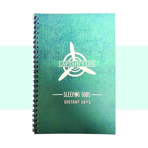May include: A spiral-bound notebook with a teal and green textured cover. The cover features a white propeller design and the text "CAPTAIN'S LOG" above "SLEEPING GODS DISTANT SKYS". The notebook has a teal spine.
