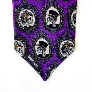May include: Purple bandana with a repeating pattern of ornate frames containing black and white skull illustrations. The bandana has the text "bitter rosa" printed on the bottom corner.