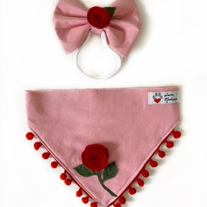 Może przedstawiać: R&oacute;żowa bandana dla psa z czerwoną ozdobą z pompon&oacute;w i czerwoną r&oacute;żą z filcu. Bandana ma białą etykietę z tekstem "Love, Matisse" i czerwonym sercem.