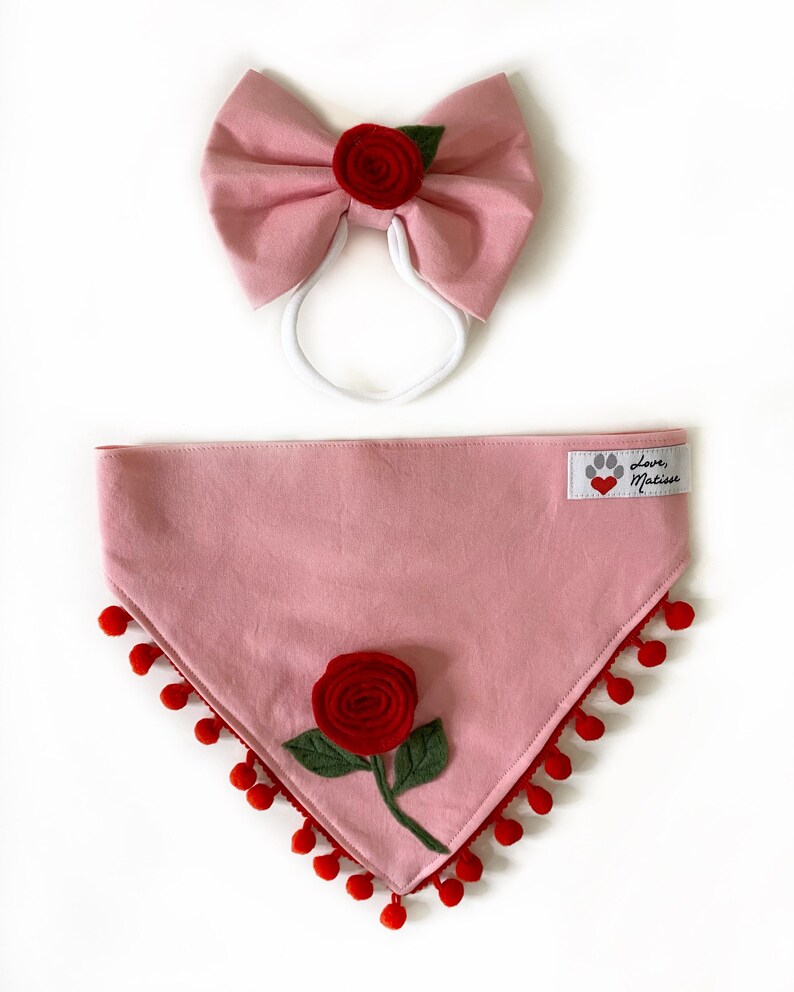 Może przedstawiać: R&oacute;żowa bandana dla psa z czerwoną ozdobą z pompon&oacute;w i czerwoną r&oacute;żą z filcu. Bandana ma białą etykietę z tekstem "Love, Matisse" i czerwonym sercem.