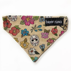 May include: A triangular pet bandana in a tan fabric featuring a colorful floral pattern interspersed with white hockey masks. A black rectangular fabric label with the white text bitter rosa is sewn onto the upper right corner of the bandana.