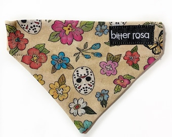 Puede incluir: Un pañuelo triangular para mascotas de tela color canela con un estampado floral colorido intercalado con máscaras de hockey blancas. Una etiqueta de tela rectangular negra con el texto blanco bitter rosa está cosida en la esquina superior derecha.