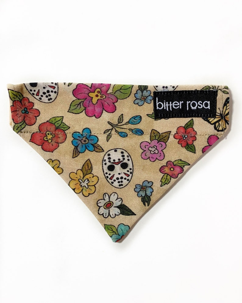May include: A triangular pet bandana in a tan fabric featuring a colorful floral pattern interspersed with white hockey masks. A black rectangular fabric label with the white text bitter rosa is sewn onto the upper right corner of the bandana.