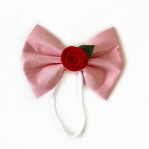 Puede incluir: Un lazo de tela rosa con una rosa de fieltro rojo y una hoja de fieltro verde en el centro. El lazo está unido a una diadema elástica blanca.