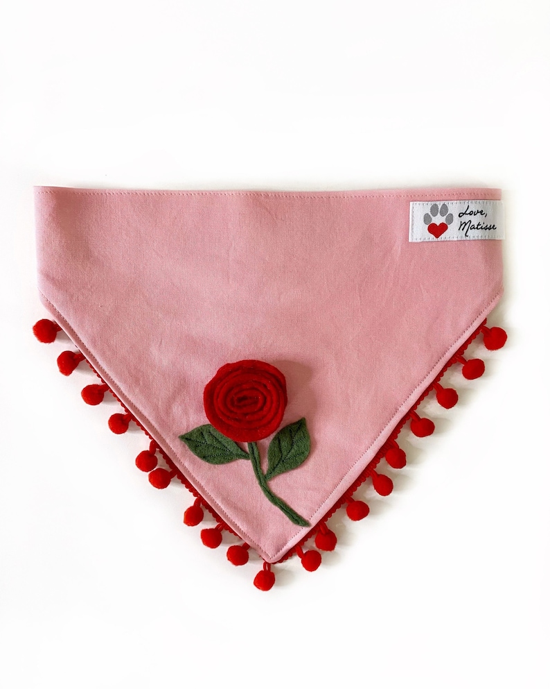 Może przedstawiać: R&oacute;żowa bandana dla psa z czerwoną filcową r&oacute;żą i czerwoną ozdobą z pompon&oacute;w. Bandana ma białą etykietę z tekstem "Love, Matisse" i czerwonym sercem.