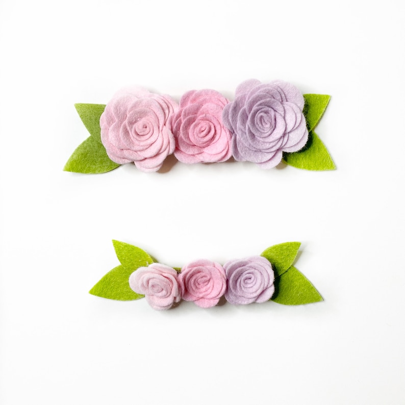 Puede incluir: Dos arreglos florales de fieltro. Cada arreglo presenta tres rosas de colores pastel en tonos rosa y lavanda, con hojas de fieltro verde. Los arreglos est&aacute;n sobre un fondo blanco.