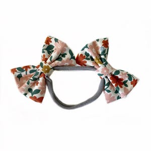 May include: A floral headband with two large bows. The bows are made of a white fabric with a pink, orange, and green floral pattern. The headband is grey.