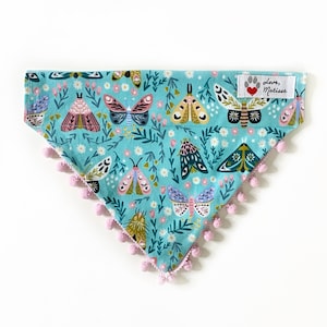 Op de afbeelding: Een driehoekige bandana voor huisdieren in lichtblauw met een herhalend patroon van kleurrijke motten, kleine bloemen en groen gebladerte. De onderrand is afgewerkt met kleine lichtroze pompons. Een wit stoffen label toont Love, Matisse.