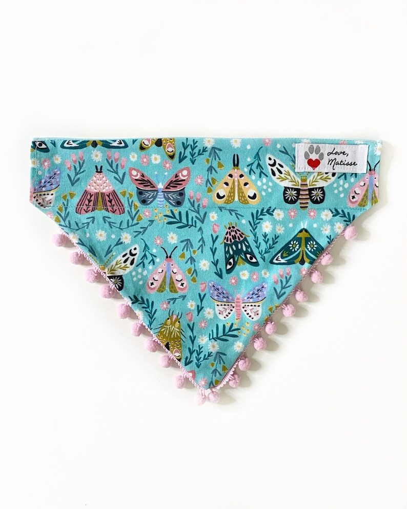 Op de afbeelding: Een driehoekige bandana voor huisdieren in lichtblauw met een herhalend patroon van kleurrijke motten, kleine bloemen en groen gebladerte. De onderrand is afgewerkt met kleine lichtroze pompons. Een wit stoffen label toont Love, Matisse.