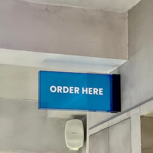 May include: A blue rectangular sign with the words "ORDER HERE" in white lettering is mounted on a gray wall. The sign is illuminated and positioned above a white speaker and a security camera.