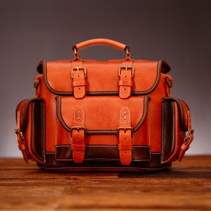May include: A leather backpack in shades of orange and brown, featuring multiple compartments and secure buckle closures. The bag has a top handle and side pockets, ideal for carrying essentials. The bag is set on a wooden surface.
