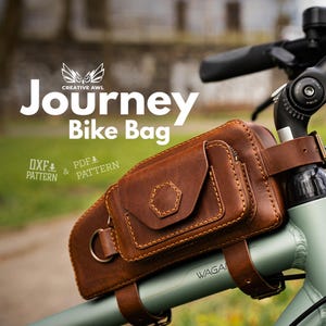 Op de afbeelding: Een bruine leren Journey fietstas bevestigd aan een fiets. De tas heeft een klepsluiting en een klein vak. De tekst "Journey Bike Bag" wordt in het wit weergegeven, samen met "DXF & PDF PATTERN". De fiets is lichtgroen.