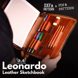 May include: A brown leather sketchbook, open to show a page with a grid pattern. Inside are pen and pencil holders, containing pens, pencils, and an eraser. The text "Leonardo Leather Sketchbook" is displayed below the sketchbook.