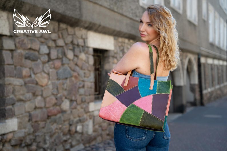 May include: A rectangular tote bag constructed from multi-coloured leather patches in shades of pink, purple, teal, green, and tan. The bag features visible decorative stitching along the seams and two leather shoulder straps in tan and green.