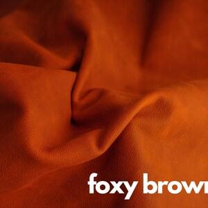 May include: Close-up of a textured, foxy brown leather fabric. The fabric is folded and draped, showcasing its soft, supple texture.