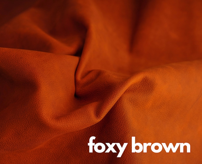 May include: Close-up of a textured, foxy brown leather fabric. The fabric is folded and draped, showcasing its soft, supple texture.