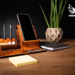 May include: A brown leather desk organizer with a phone holder, pen holders, and a small notepad. The organizer is on a wooden desk with a plant and a notebook in the background. The Creative Awl logo is in the top right corner.