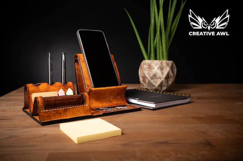 May include: A brown leather desk organizer with a phone holder, pen holders, and a small notepad. The organizer is on a wooden desk with a plant and a notebook in the background. The Creative Awl logo is in the top right corner.