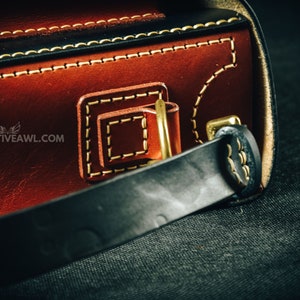 May include: Close-up of a handcrafted leather bag with intricate stitching and brass hardware. The bag features a rich brown exterior with black accents and a sturdy black strap. The website creativeawl.com is visible.