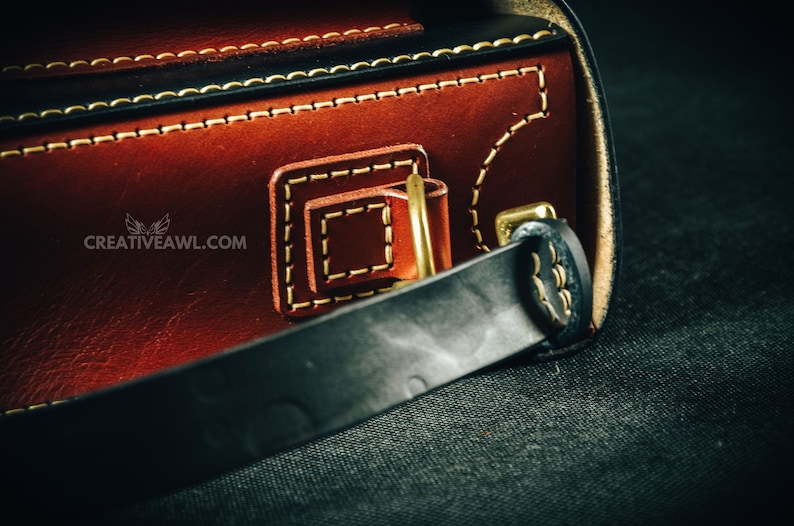 May include: Close-up of a handcrafted leather bag with intricate stitching and brass hardware. The bag features a rich brown exterior with black accents and a sturdy black strap. The website creativeawl.com is visible.
